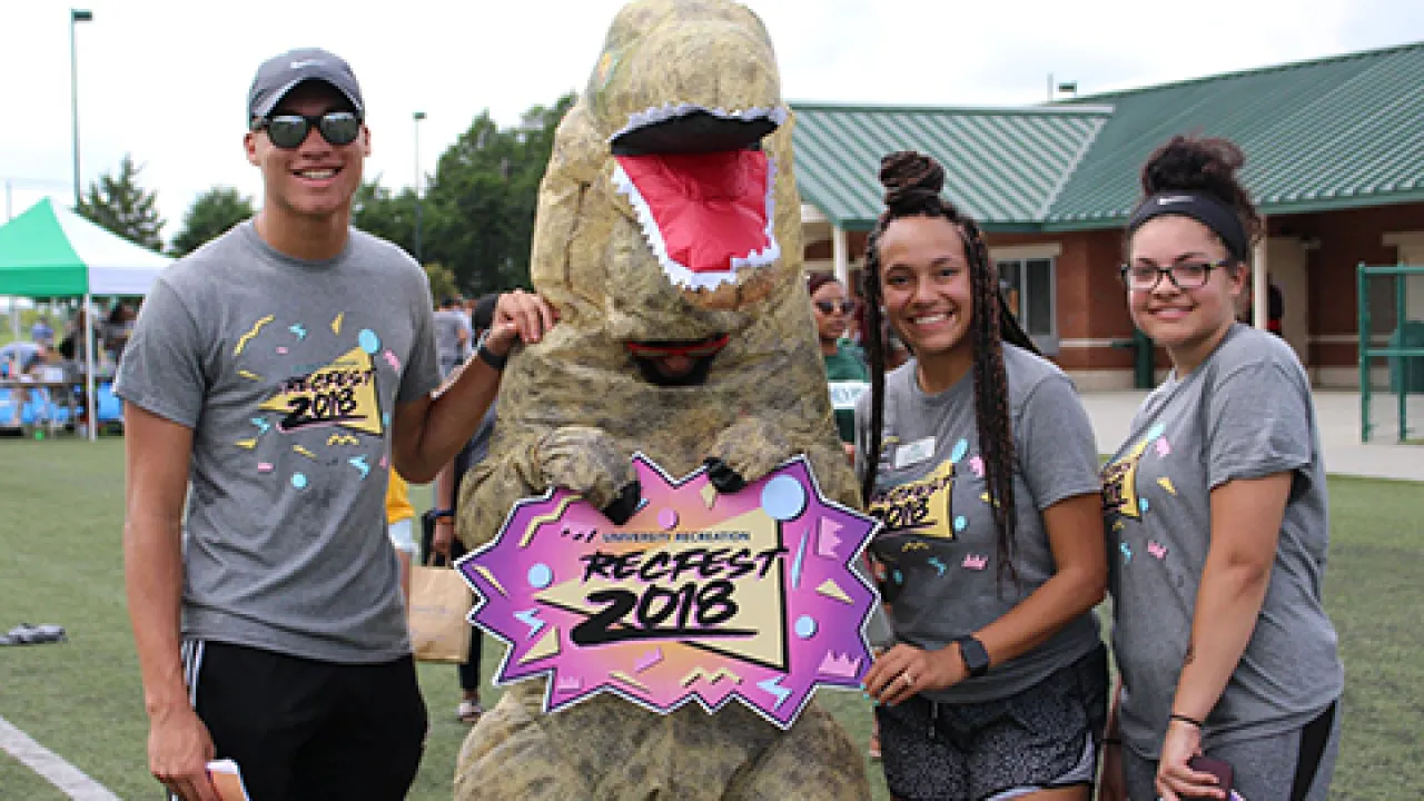 3 people standing next to a dinosaur holding a sign that reads RECFEST 2018.